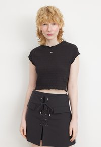 Sort cropped top med smocket tekstur, korte cap-ærmer og bølget kant. Stylet med en højtaljet sort nederdel med snorede detaljer.