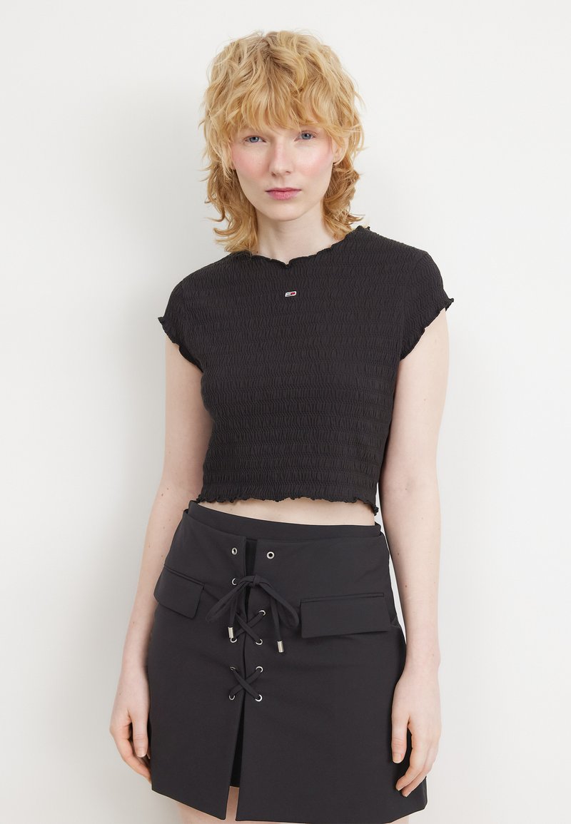 Sort cropped top med smocket tekstur, korte cap-ærmer og bølget kant. Stylet med en højtaljet sort nederdel med snorede detaljer.