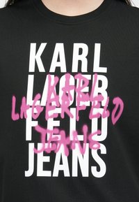 T-shirt preta com texto branco em destaque a ler "KARL LAGERFELD JEANS," sobreposto por letras em rosa spray.