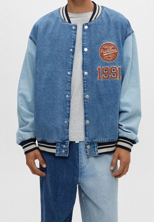 Man draagt een tweekleurig denim varsity jack met "Pull&Bear" patch en "1991" op de borst, gecombineerd met tweekleurige denim jeans.