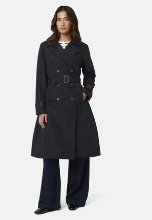 Vrouw draagt een lange zwarte dubbelgeknoopte trenchcoat met riem, brede donkere jeans en zwarte schoenen, staand met één hand in de zak.