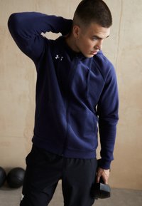 Marineblauwe zip-up hoodie met een getailleerd design, gemaakt van een katoenmix, met twee voorzakken en een capuchon met trekkoord. Model houdt een dumbbell vast.