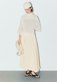 Beige gestricktes Oberteil mit kurzen Ärmeln, kombiniert mit einem langen, hellgelben Rock. Schwarze Sandalen und eine beige Tasche runden das Outfit ab.