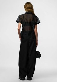 Haut court noir transparent à manches courtes avec un corsage ajusté, associé à un pantalon large noir taille haute. Un petit sac à main noir pend sur le côté.