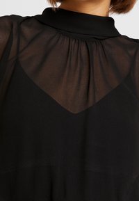 Mustat läpinäkyvät toppi korkean kaula-aukolla, laskostetuin kankaan yksityiskohdin ja V-muotoisella suunnittelulla. Materiaali näyttää kevyeltä ja läpinäkyvältä.