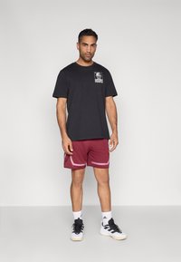 Zwart katoenen T-shirt met grafische opdruk, bordeauxrode sportbroek met lichtroze zijaccenten, en zwarte sportschoenen met witte zolen.