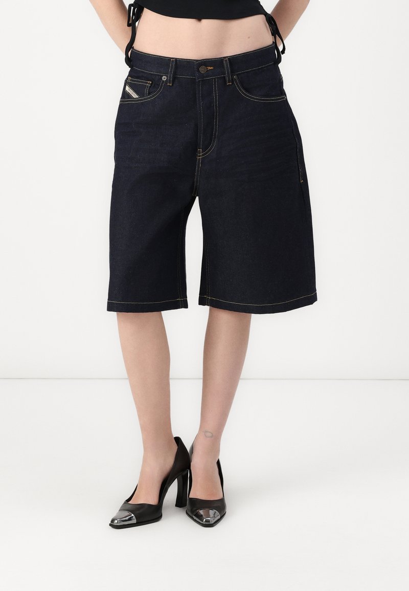 Jeans bermuda hlače v temno modri barvi z rumenimi šivi, ki imajo sprednje žepke in ohlapen kroj. V kombinaciji s črnimi visokimi pete.