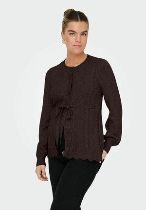 Femme portant un cardigan à torsades marron foncé avec ceinture à nouer, sur un pantalon noir, debout devant un fond clair uni.