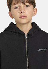 Sudadera negra con cremallera hecha de un tejido suave, con una cremallera plateada y un pequeño logo gris en el pecho. Diseño simple con capucha.