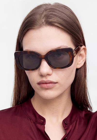 Jeune femme aux cheveux bruns raides, portant de grandes lunettes de soleil écaille foncées et un haut bordeaux boutonné sur un fond uni.