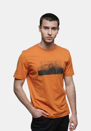 Giovane uomo con capelli corti e scuri che indossa una maglietta arancione con una grafica nera e un disegno a codice a barre, in piedi con una mano in tasca.
