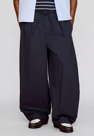 Muž nosí voľné tmavomodré široké nohavice so šnúrkou v páse, biele a čierne mokasíny a svetlomodrú košeľu s krátkym rukávom.