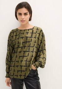 Blusa verde oliva con un motivo a griglia in pennellate nere, scollatura rotonda, vestibilità ampia e maniche arricciate; abbinata a pantaloni neri.