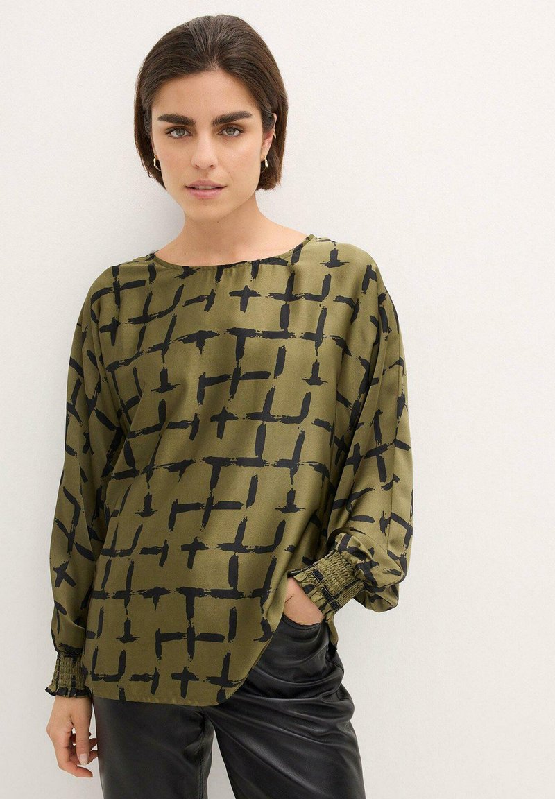 Blusa verde oliva con un motivo a griglia in pennellate nere, scollatura rotonda, vestibilità ampia e maniche arricciate; abbinata a pantaloni neri.