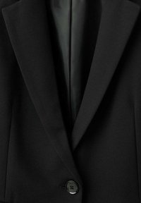 Zbliżenie na czarną dopasowaną marynarkę z wyciętymi klapami, pojedynczym czarnym guzikiem i gładką podszewką widoczną w centrum.