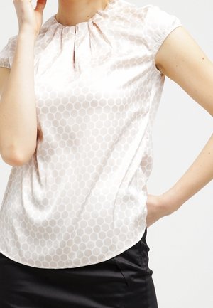 Woman wearing a short-sleeve cream blouse with a subtle circular pattern and black skirt, posing with one hand on her hip.