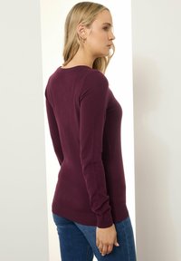 Bordeaux lange mouwen sweater gemaakt van zachte stof, met een ronde hals en geribbelde zoom. Gedragen met blauwe denim jeans.