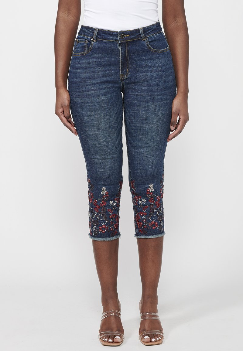 Dark denim capri jeans with floral embroidery in red and white at the hem, featuring a mid-rise waist and five-pocket design.