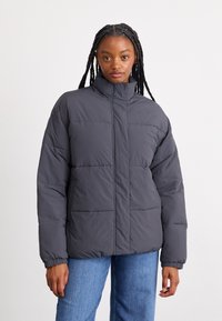 Doudoune grise à col haut, avec un design matelassé et des poignets élastiques, associée à un jean en denim bleu.