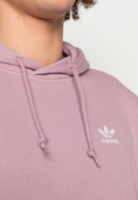 Svetle růžová mikina s kapucí z jemné látky má šňůrky na stažení kapuce a malé bílé logo Adidas. Textura působí hladce a pohodlně.
