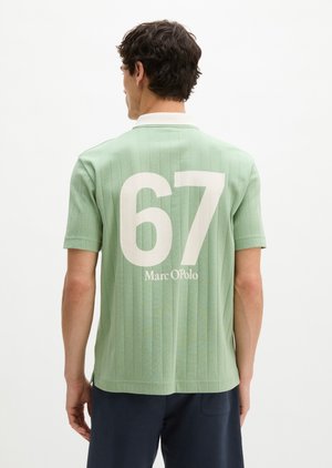 Homme portant un polo vert clair à manches courtes avec les inscriptions blanches "67" et "Marc O'Polo" dans le dos, associé à un pantalon foncé, de dos.