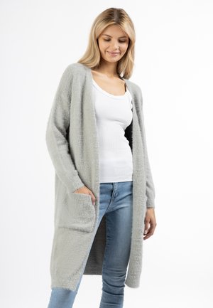 Blonde Frau trägt eine lange graue flauschige Strickjacke, ein weißes Tanktop und blaue Jeans und steht mit einer Hand in der Strickjackentasche.