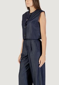 Completo in denim scuro composto da due pezzi, con top senza maniche e bottoni e pantaloni a gamba larga con pieghe laterali, che offrono un silhouette strutturata.