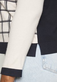 Close-up of a person's arm in a cream sweater with navy cuffs and a side slit, wearing light blue denim jeans with a visible back pocket.