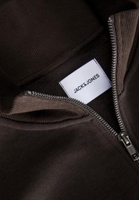 Sudadera marrón con cremallera, que presenta un cuello acanalado y detalle de cremallera. La etiqueta interior dice "JACK & JONES". Se puede ver la textura suave de la tela.