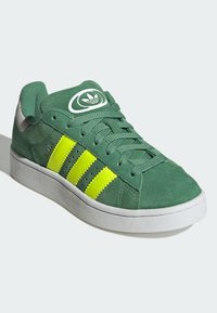 Zapatillas de ante verde con tres rayas de neón amarillo, suela de goma blanca y logo en la lengüeta. Presenta cordones texturizados y un collar acolchado.