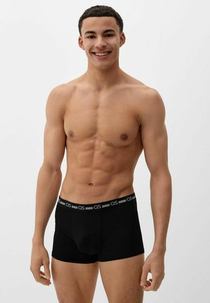Jonge man die glimlacht, gekleed in zwarte QS-merkkorte boxershorts, staand tegen een effen lichte achtergrond en zijn gespierde bovenlichaam tonend.