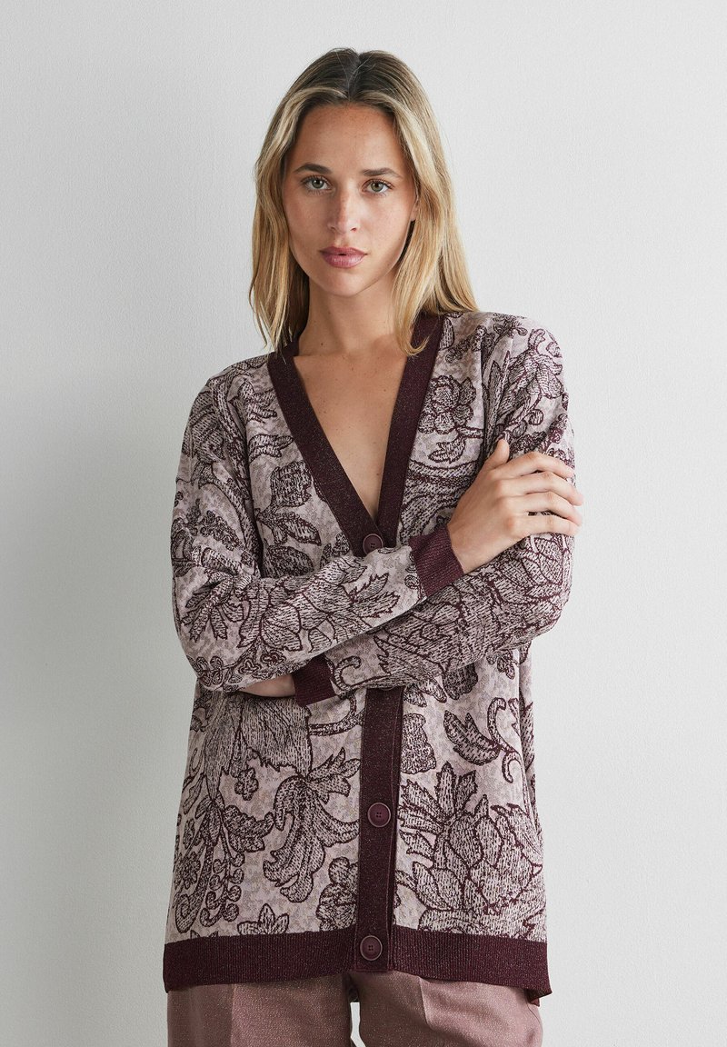 Vrouw met blond haar die een gebuttonde cardigan met bloemmotief draagt in tinten van mauve en bordeaux, staand tegen een effen witte muur.