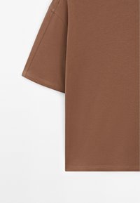 T-shirt marron en coton à manches courtes avec coutures visibles sur la manche et l’ourlet, sur fond blanc uni.