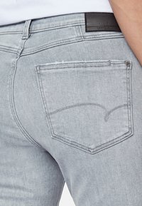 Los pantalones vaqueros de mezclilla gris claro cuentan con un bolsillo trasero con un patrón de costura decorativa y una etiqueta de cuero negro en la cintura.