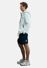 Weißer Zip-Hoodie mit schwarzem Akzentlogo, kombiniert mit blauen Shorts und silbernen Sportschuhen. Glattes Material, moderne Passform und minimalistisches Design.
