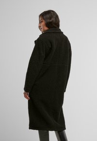 Cappotto nero, morbido, lungo, realizzato in materiale peluche con colletto e tasche laterali, caratterizzato da una vestibilità rilassata e design di lunghezza media.