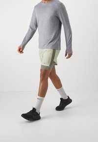 Camisa gris de manga larga combinada con pantalones cortos deportivos verdes claros con un bolsillo lateral, acompañada de calcetines blancos y zapatillas negras.