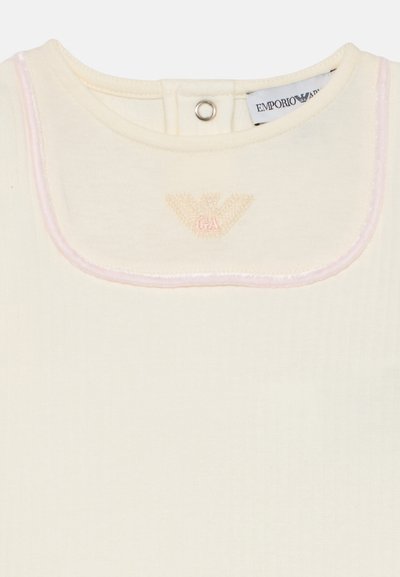 Kremfarget topp med ribbet tekstur, med en rund krage kantet i rosa sateng og en brodert logo i midten.
