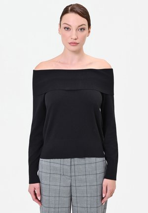 Donna con capelli castani lisci indossa una maglia nera lunga a spalle scoperte e pantaloni grigi a quadri, in piedi su sfondo bianco semplice.