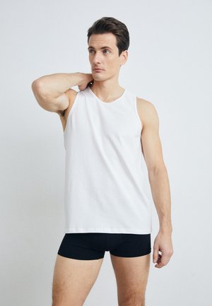White sleeveless tank top made of cotton, featuring a round neckline and relaxed fit, paired with black fitted shorts. Background is neutral.