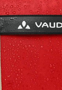 Rood waterdicht stof met zichtbare waterdruppels. Zwarte band met het witte "VAUDE" logo bovenaan. Gladde textuur met een matte afwerking.