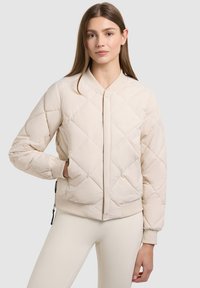 Jeune femme portant une veste bomber matelassée beige clair et un pantalon assorti, debout avec une main dans la poche de la veste.