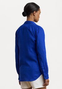 Camisa de linho azul de mangas longas com colarinho, punhos abotoados e barra curva. Textura suave, corte descontraído. Detalhe de costura visível.