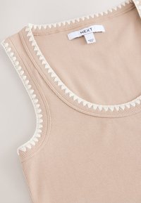Débardeur côtelé beige avec un bord festonné blanc le long de l'encolure et des emmanchures. Étiquette portant la mention "NEXT." Texture lisse et coupe ajustée.