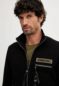 Giacca nera con zip e collo alto, dotata di una tasca con zip frontale e patch "AERONAUTICA". Indossata sopra una camicia verde oliva.