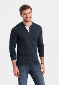 Hombre sonriente que lleva una camiseta henley de manga larga azul marino con una tapeta de botones blanca y jeans azules, de pie contra un fondo blanco liso.