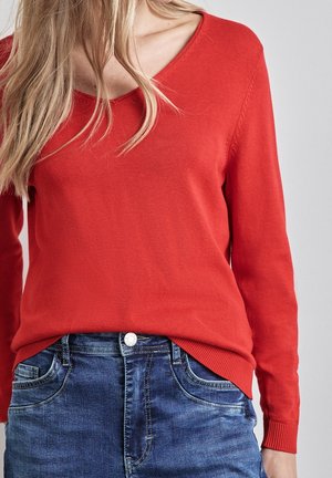 Frau trägt einen roten V-Ausschnitt-Pullover und blaue Jeans, dargestellt von den Schultern bis zur Hüfte vor einem einfarbigen Hintergrund.