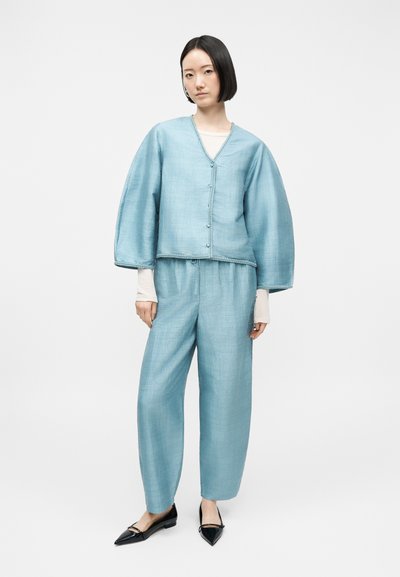 Γυναίκα που φοράει ανοιχτό μπλε λινό σακάκι και αντίστοιχο παντελόνι πάνω από λευκό μακρυμάνικο τοπ, με μαύρα μυτερά παπούτσια, στέκεται σε λευκό φόντο.