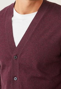 Cardigan bordeaux avec col en V, fermeture à boutons et texture douce. Comprend deux boutons foncés et un look superposé sur un t-shirt blanc.