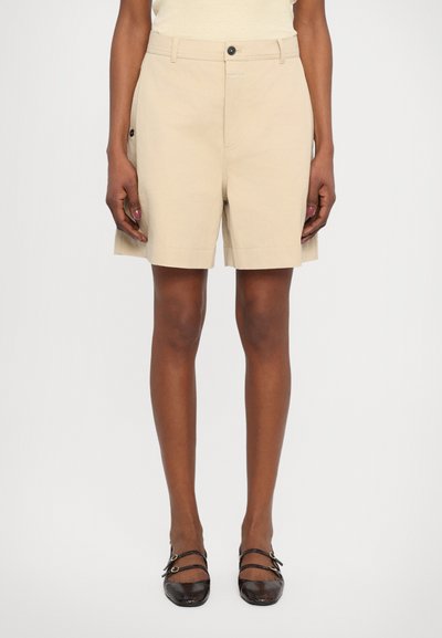 Beige cotton shorts with a straight cut, featuring a button closure, side pockets, and a clean hem. Paired with dark textured flats.
